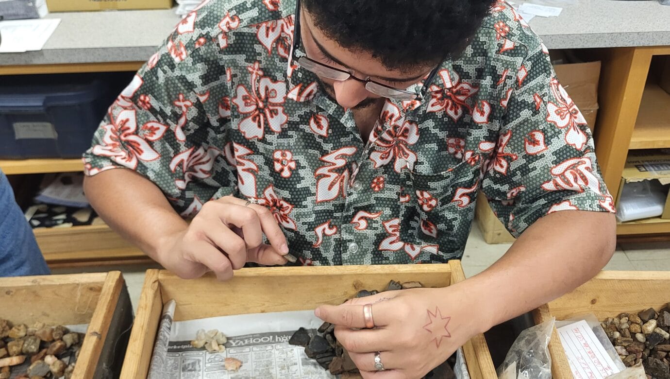 A young man in glasses and a floral shirt sorts small stones and pottery fragments in a wooden tray lined with newspaper.